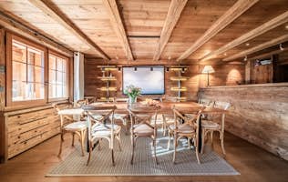 Verbier - 3 Verbier accommodation - Apartment Snowcrest - A rustic dining room with a wooden table and chairs, a bouquet of flowers, and a wall-mounted TV. Wooden walls and ceiling, with windows on the left. A carpet is under the table.
