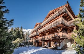 Verbier - 6 Verbier accommodation - Apartment Snowcrest - A wooden alpine chalet with balconies is surrounded by snow and trees, set against a clear blue sky and mountain backdrop.