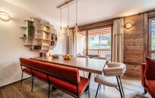 Les Gets accommodation - Apartment Colibri - Modern dining room with a wooden table, red chairs, pendant lights, and a shelf, featuring large windows with a view of greenery.