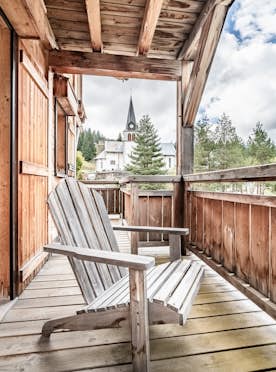 Les Gets - 5 Chamonix alojamiento - Apartamento Solstice - Wooden balcony with two Adirondack chairs, wooden railings, and a view of trees and a church with a tall steeple in the background under a partly cloudy sky.