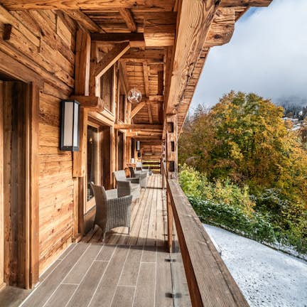 Wooden balcony of a rustic cabin with a view of snowy trees and a foggy mountain landscape, featuring outdoor furniture and a glass wall.