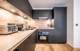 Les Gets - 6 Chamonix alojamiento - Apartamento Dolmen - Modern kitchen with black cabinets, stainless steel appliances, and wooden countertops. Under-cabinet lighting illuminates the workspace. A dining table is partially visible on the left.