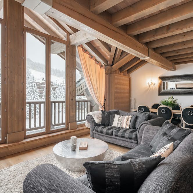 Verbier alojamiento - Apartamento Aelora - Beautiful open plan dining room at ski in ski out apartment Itauba Courchevel 1850 Verbier alojamiento - Apartamento Aelora - Cozy living room with wooden beams, large windows showing a snowy landscape, gray sofas, a round coffee table, and a dining area.