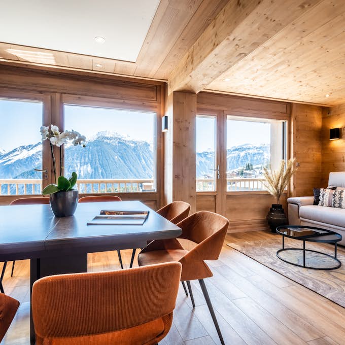 A cozy, wooden-finished living room with large windows offering a mountain view. The room includes a table with orange chairs, a sofa, and a plant.