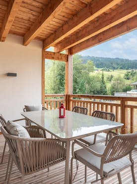 Praz sur Arly - 5 Praz-sur-Arly alojamiento - Apartamento Tavoro - A covered wooden patio with a glass dining table, six chairs, and a red lantern centerpiece, overlooking neighboring houses and distant hills under a partly cloudy sky.