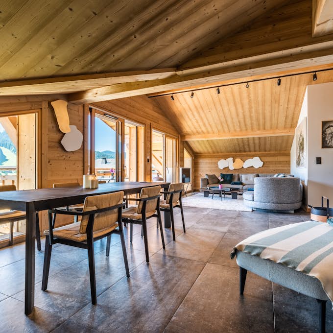 Service de conciergerie à Les Gets  Conciergerie Les Gets Spacious open-plan living and dining area with wooden ceiling and large windows offering mountain views. Modern furnishings include a dining table, chairs, a gray couch, and a side table with a lamp.