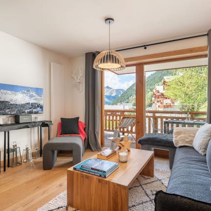 Apartment Tilia, Champagny-en-Vanoise Cozy living room with a gray sofa, wooden coffee table, TV, and large window revealing a scenic mountain view. Modern decor and warm lighting.