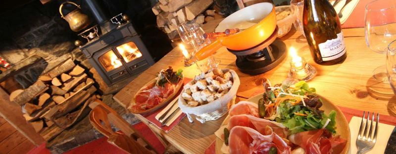 A rustic dining setup featuring a fondue pot, two plates with charcuterie and salad, a lit candle, wine bottle, and a fireplace with stacked wood in the background. A rustic dining setup featuring a fondue pot, two plates with charcuterie and salad, a lit candle, wine bottle, and a fireplace with stacked wood in the background.