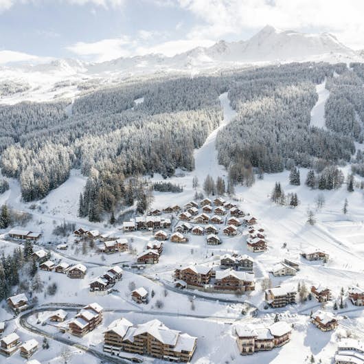 A snow-covered alpine village nestled in a mountainous landscape with houses, trees, and winding roads below a backdrop of cloudy sky and peaks.