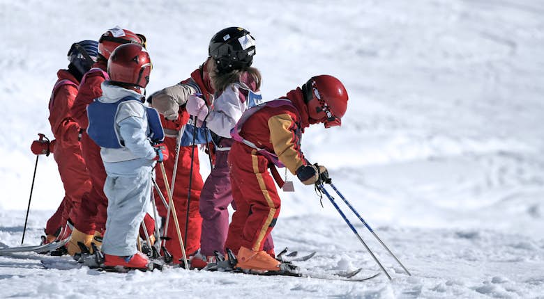 A group of children in skiing gear are learning to ski on a snowy slope. They are dressed in colorful outfits and helmets, standing in a line with their skis on.
