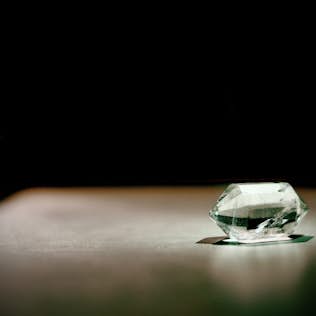 A large, clear cut gemstone sits in focus on a smooth surface, illuminated by a spotlight in a dimly lit setting.