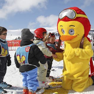 A mascot dressed as a yellow bird with a helmet and goggles interacts with children in ski gear on a snowy hill.