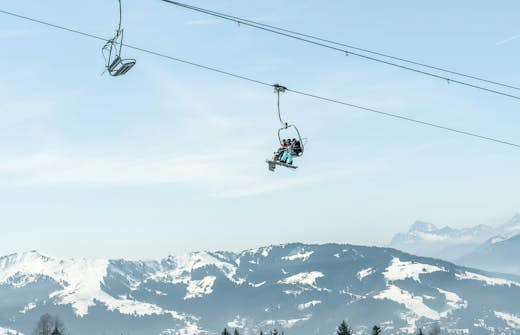 Two people on a ski lift over snowy mountains with clear blue skies.