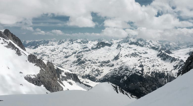 A panoramic view of snow-covered mountains under a partly cloudy sky. Rocky ridges and snow-capped peaks stretch into the distance.