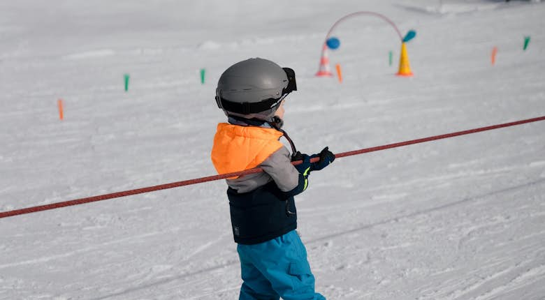 A child dressed in a helmet, orange vest, and blue pants skis while holding onto a red rope tow on a snowy slope with ski training aids in the background.