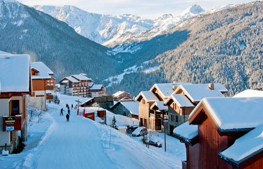 Snow-covered village with mountain backdrop, people walking on snow-covered roads, and red-roofed buildings bathed in sunlight.