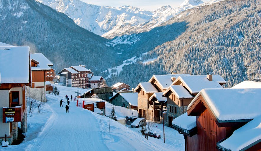 Snow-covered village with mountain backdrop, people walking on snow-covered roads, and red-roofed buildings bathed in sunlight.