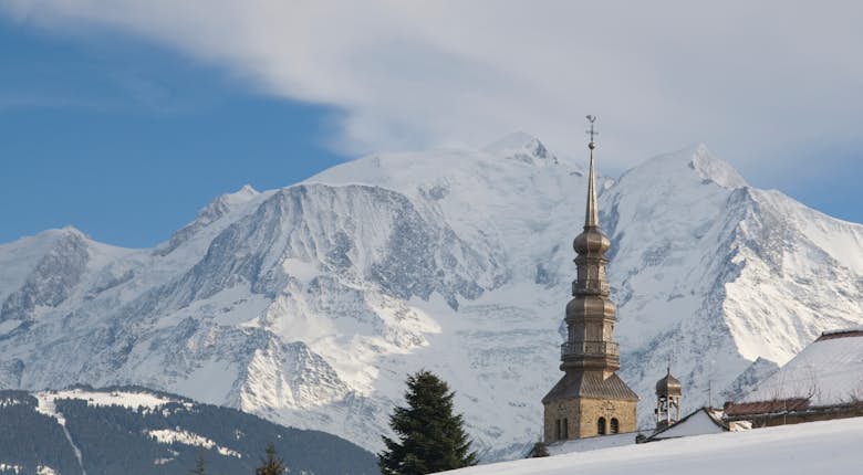 A snow-covered landscape with a church steeple in the foreground, against the backdrop of majestic, snow-capped mountains under a partly cloudy sky.