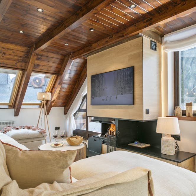 Val de Ruda - 1 Baqueira Beret accommodation - Apartment Nevada - Modern living room with light wood ceiling beams, large windows, a gray sectional sofa, a wooden coffee table, poufs, and a fireplace with a TV mounted next to it. Scenic mountains are visible outside.