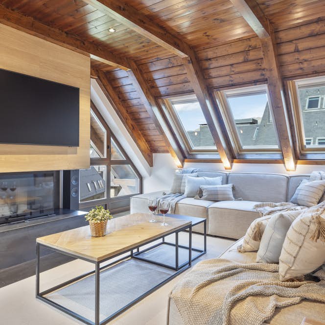 Val de Ruda - 1 Baqueira Beret location - Appartement Galena - Modern living room with light wood ceiling beams, large windows, a gray sectional sofa, a wooden coffee table, poufs, and a fireplace with a TV mounted next to it. Scenic mountains are visible outside.