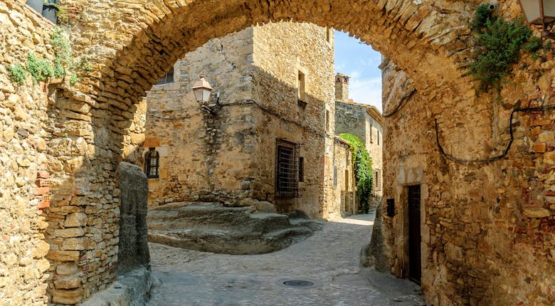 Stone archway and cobblestone street in a historic village with old buildings and a lamp mounted on the wall under clear blue sky.