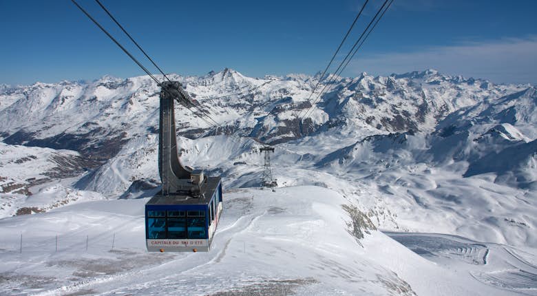 A cable car travels through snowy mountainous terrain under a clear blue sky, suspended by cables attached to tall supporting towers.