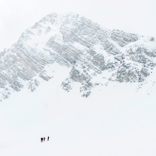 Four people are climbing a snowy mountain with steep, jagged rock face under a cloudy sky.