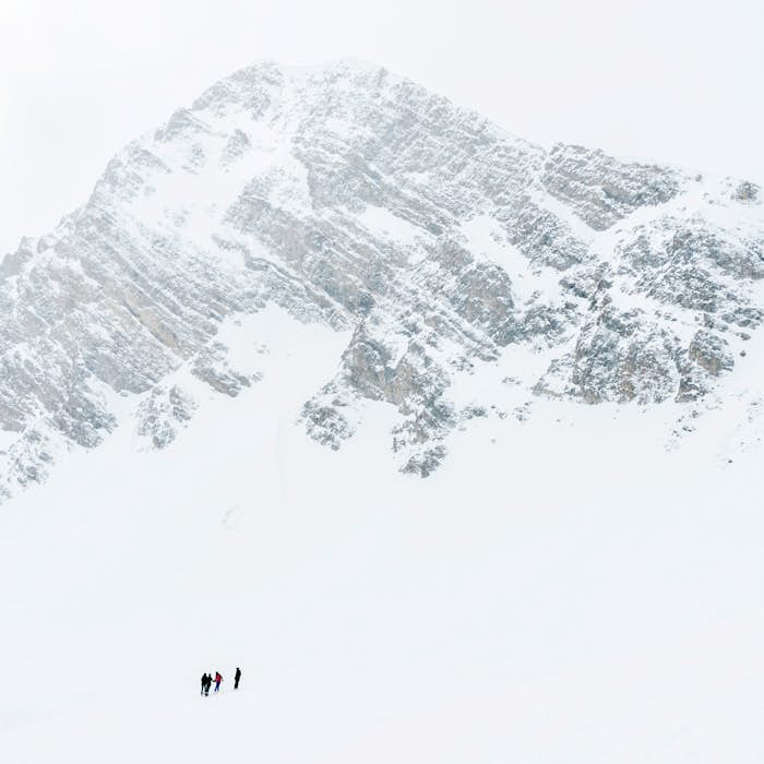 Four people are climbing a snowy mountain with steep, jagged rock face under a cloudy sky.