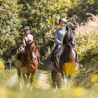 Two women riding horses on a sunny forest trail, smiling and wearing helmets. flowers in the foreground.