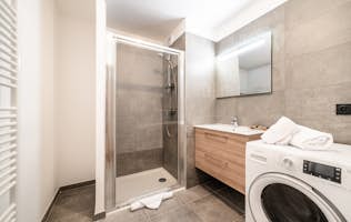 Les Gets accommodation - Apartment Primerose - Modern bathroom with a glass shower, wooden vanity, and washing machine. Walls and floor are tiled in gray. A mirror is above the sink. Towels are folded on the washing machine. Les Gets accommodation - Apartment Primerose - Modern bathroom with a glass shower, wooden vanity, and washing machine. Walls and floor are tiled in gray. A mirror is above the sink. Towels are folded on the washing machine.