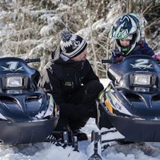 A man and a young child wearing helmets sit on parked snowmobiles in a snowy forest, looking at each other and talking.