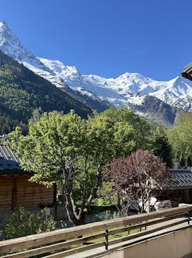 Chamonix - 5 Chamonix accommodation - Chalet Inari - A view from a balcony showing snowy mountains, green trees, and wooden houses under a clear blue sky.