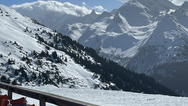 Snow-covered mountains under a clear sky, with a wooden deck in the foreground set with red chairs and tables for dining.
