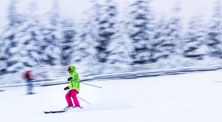A person wearing bright green and pink ski gear is skiing down a snow-covered slope with blurred snow-covered trees in the background.