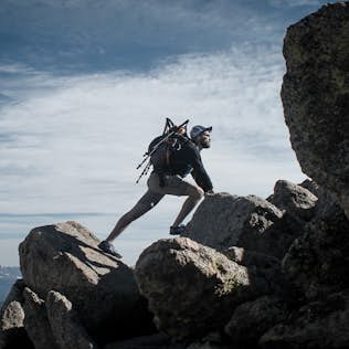 Hiker climbing over rocks with a backpack in mountainous terrain under a clear blue sky.