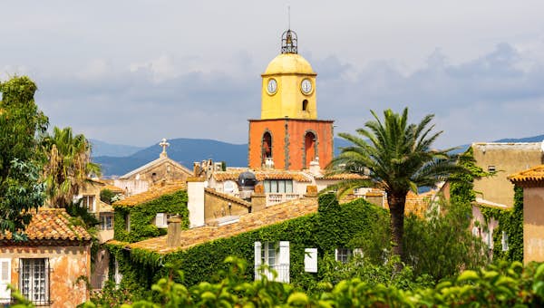 Image by xbrchx on Adobe Stock View of a coastal town with red-tiled roofs, a prominent clock tower, and a body of water in the background, framed by a rocky window opening.