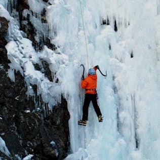 A climber in orange and blue gear ascends a frozen waterfall using ice axes and crampons, surrounded by icicles.