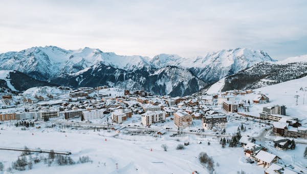 Mountain view from a wooden balcony, overlooking snowy peaks and a skier below under a cloudy sky.