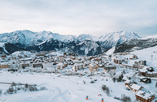 Mountain view from a wooden balcony, overlooking snowy peaks and a skier below under a cloudy sky.