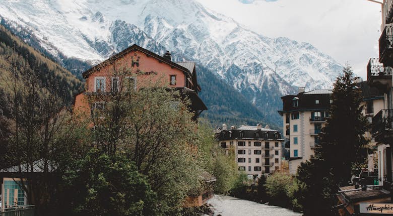 A small river runs through a picturesque alpine town with snow-capped mountains in the background. The town features buildings with balconies and a mix of trees and greenery.