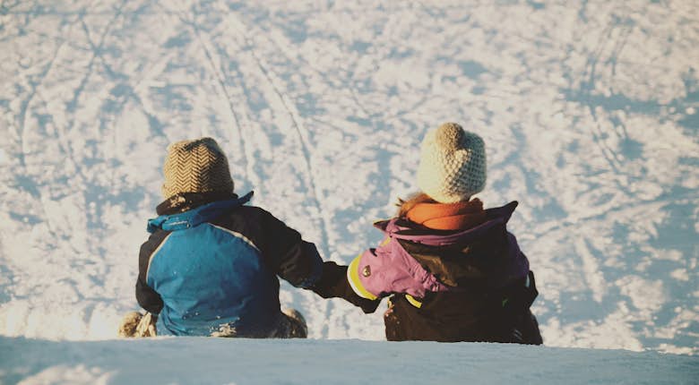 Two children, dressed in winter jackets and knit hats, sit in the snow holding hands, facing away from the camera.