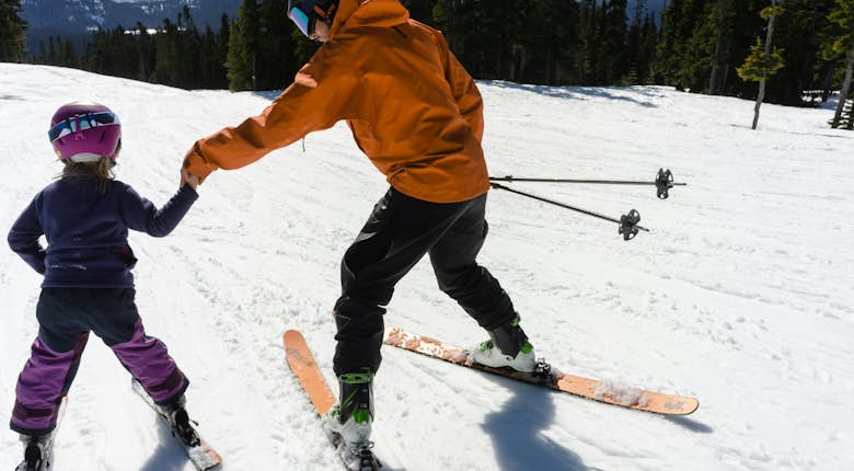 Two children wearing ski gear and helmets smile while standing outside a ski lodge labeled