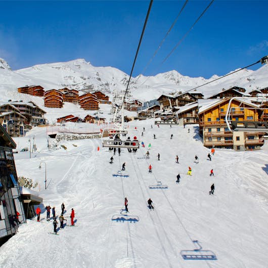 A snowy ski resort scene with skiers on the slopes, ski lifts in motion, and mountain lodges in the background under a clear blue sky.