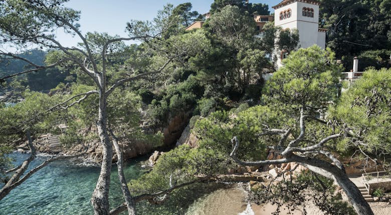 Scenic coastal view featuring a sandy cove, clear blue water, and rugged trees with a traditional mediterranean-style villa nestled among green foliage in the background.