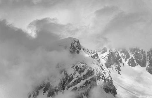 Snow-covered mountain peak shrouded by swirling clouds in a grayscale image.