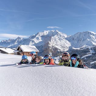 Group of skiers smiling and peering over a snowbank, with mountains and a cabin in the background on a sunny day.