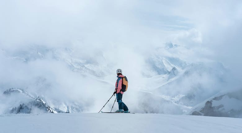 Skier in a pink jacket stands on a snowy mountain slope with misty clouds and distant snow-covered peaks in the background.