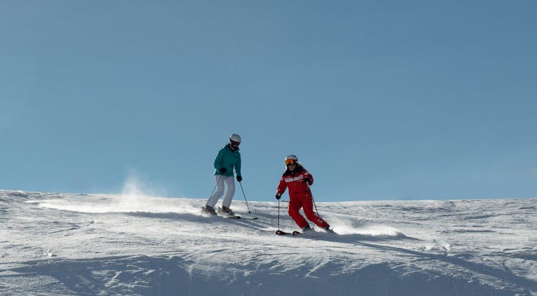 Two skiers in colorful gear glide down a snowy slope, with clear blue skies in the background.