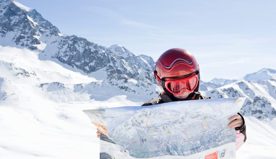 A skier in a helmet and goggles holds a map in front of snowy mountain peaks.
