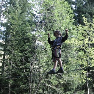 A person wearing a helmet and harness walking on a rope high up among tall green trees, using guide wires for balance.